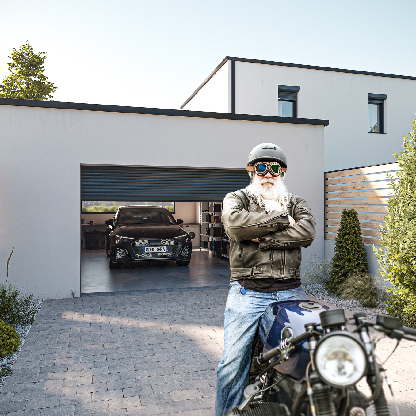 Mann mit weißem Bart sitzt in einer Auffahrt auf einem Motorrad, dahinter eine Garage mit halb heruntergelassenem Garagentor und einem darin parkenden Auto.